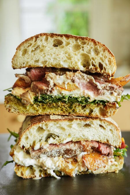 A close-up of a stacked sandwich cut in half, showing layers of sliced steak, creamy cheese, leafy greens, pesto, and toasted rustic bread. The sandwich sits on a dark surface with a blurred background.