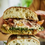 A close-up of a large sandwich cut in half, showing layers of leafy greens, sliced meat, melted cheese, sauce, and crusty artisan bread, with a person holding one half.