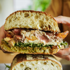 A close-up of a large sandwich cut in half, showing layers of leafy greens, sliced meat, melted cheese, sauce, and crusty artisan bread, with a person holding one half.