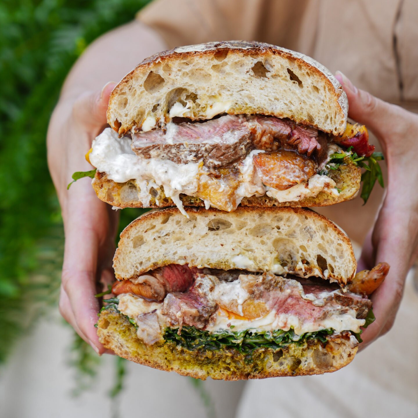 A person holds a large sandwich cut in half, filled with steak, greens, melted cheese, and a creamy sauce, all between thick slices of rustic bread.