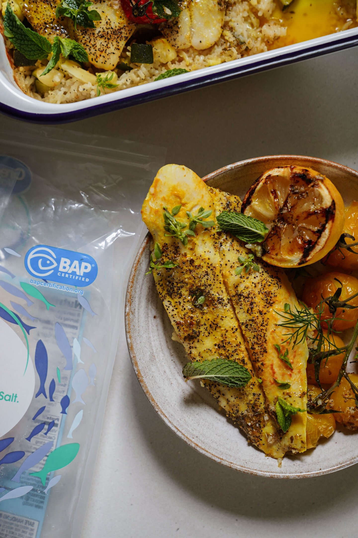A plate with seasoned, cooked fish fillets garnished with fresh herbs and grilled lemon, served alongside yellow tomatoes. Nearby is a BAP-certified seafood packaging on a white surface.