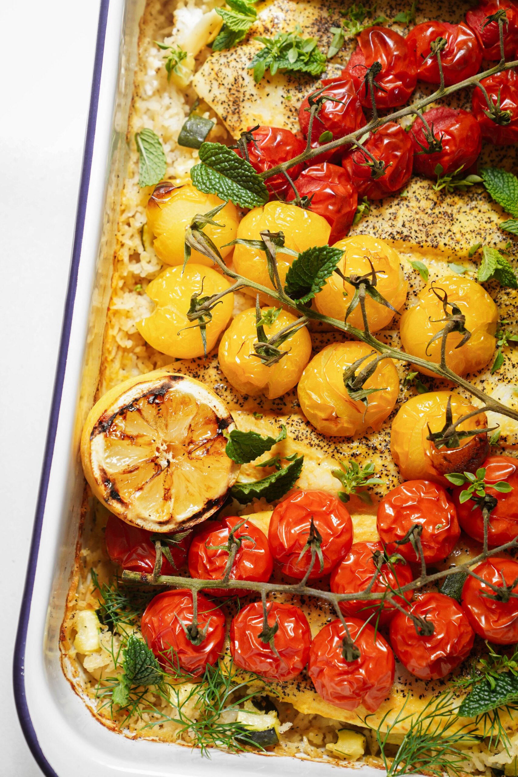 A baked dish with roasted red and yellow vine tomatoes, a halved grilled lemon, herbs, and seasoned white fish or chicken in a white enamel baking tray.