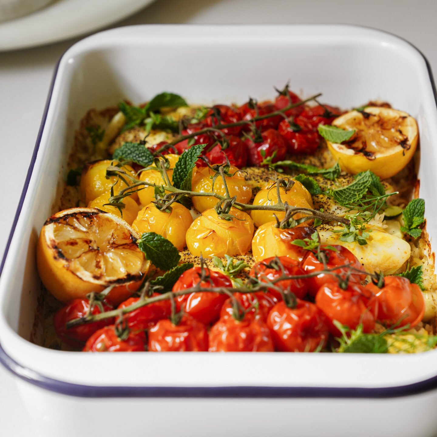 A white baking dish filled with roasted red and yellow tomatoes on the vine, charred lemon halves, fresh mint leaves, and herbs, arranged in colorful rows.
