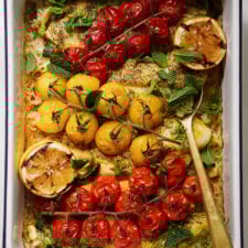 A baked dish with roasted red and yellow tomatoes on the vine, grilled lemon halves, fresh mint and dill, and a golden serving spoon on the side. The vegetables are arranged on a bed of what appears to be cheese or yogurt.