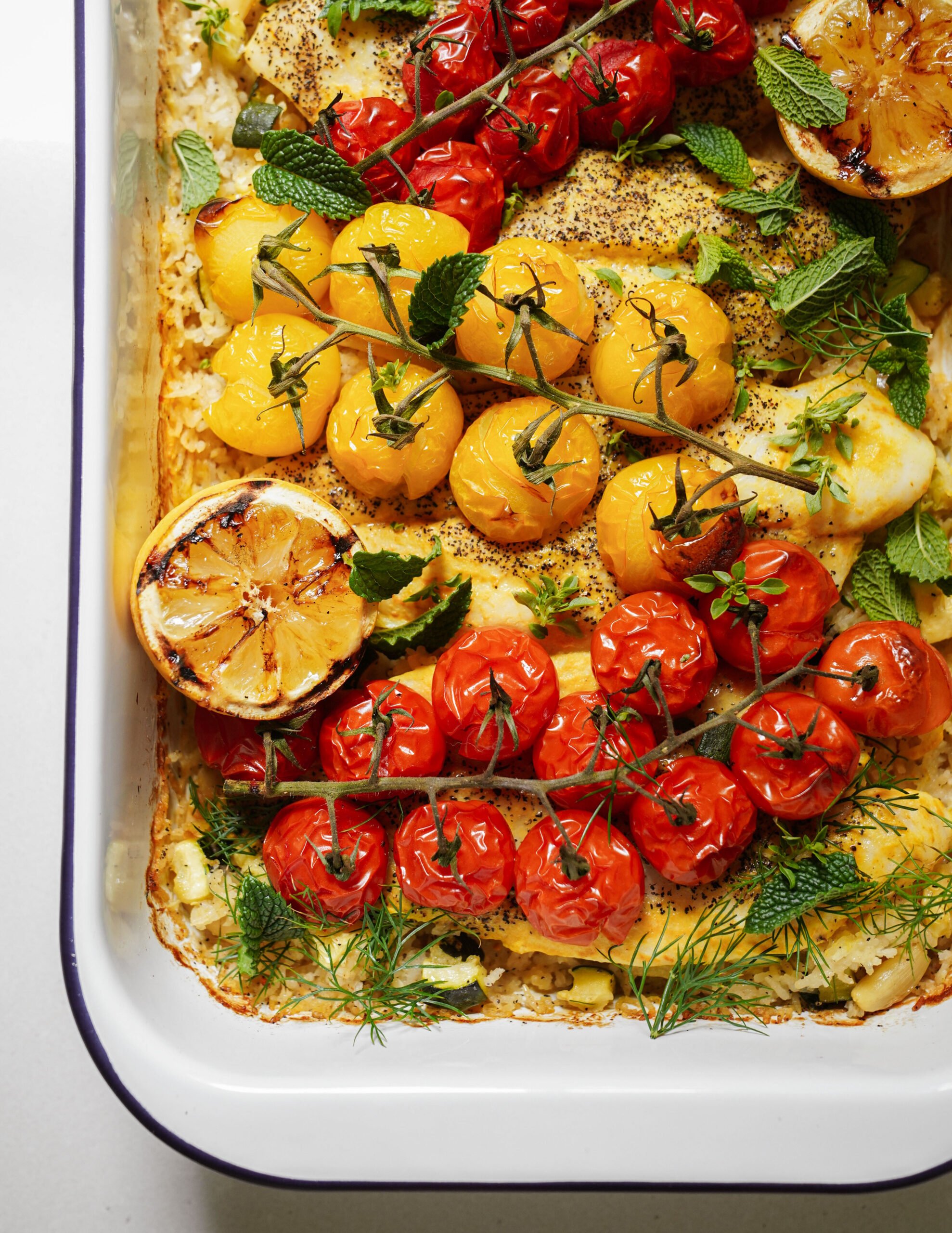 A baked dish topped with roasted yellow and red cherry tomatoes on the vine, fresh herbs, and halved grilled lemons, displayed in a white rectangular baking dish.