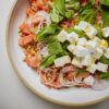 A close-up of a fresh salad with diced watermelon, feta cheese cubes, sliced red onions, chopped pistachios, basil, and mint leaves on a white plate.