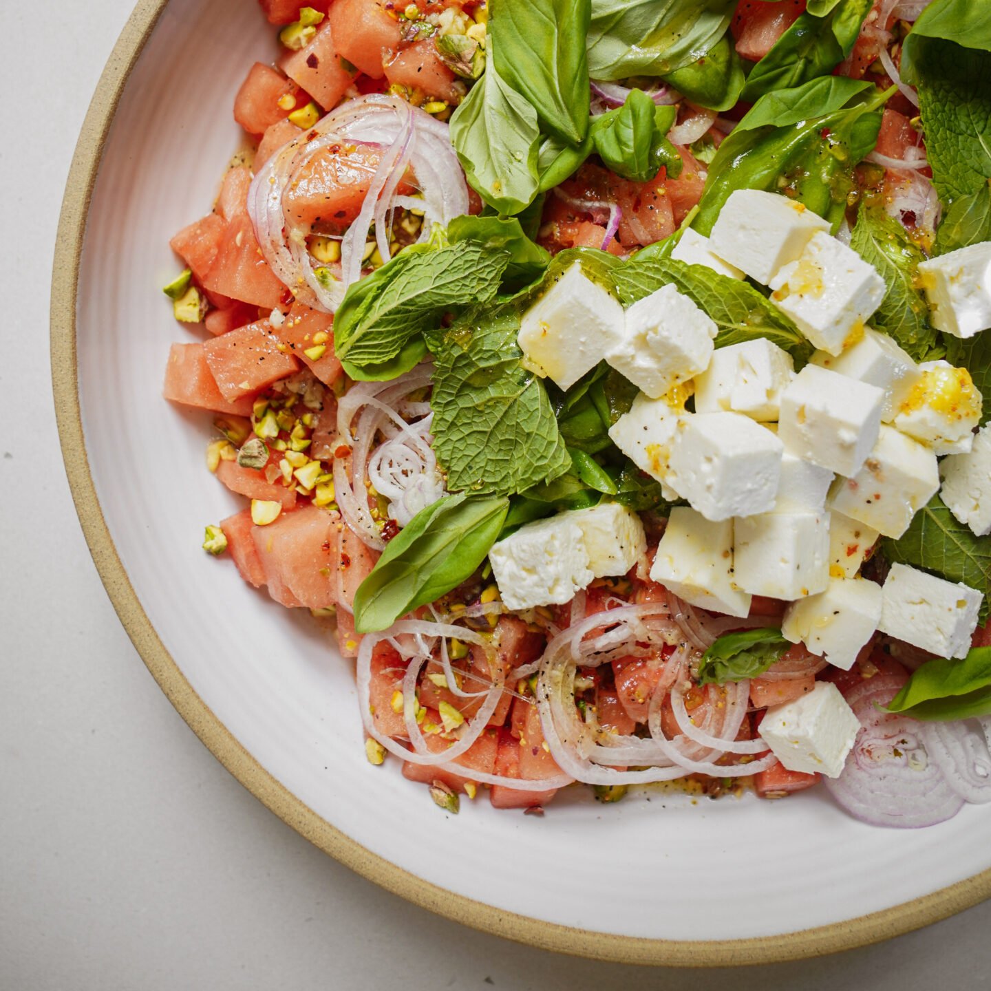 A close-up of a fresh salad with diced watermelon, feta cheese cubes, sliced red onions, chopped pistachios, basil, and mint leaves on a white plate.