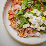 A close-up of a fresh salad with diced watermelon, feta cheese cubes, sliced red onions, chopped pistachios, basil, and mint leaves on a white plate.