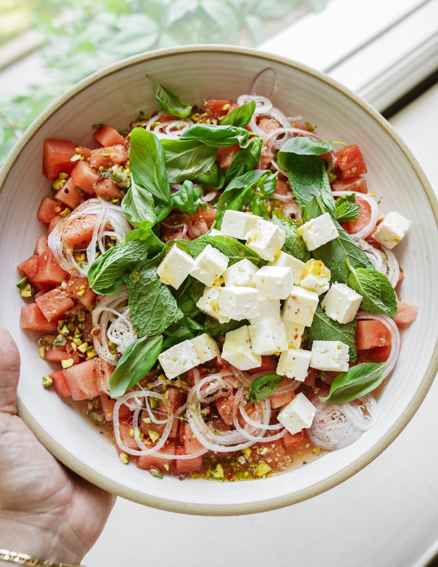 A hand holds a bowl of salad with diced watermelon, sliced onions, feta cheese cubes, fresh mint and basil leaves, and chopped pistachios, set near a window with greenery visible outside.