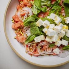 A close-up of a fresh salad with diced watermelon, feta cheese cubes, sliced red onions, chopped pistachios, basil, and mint leaves on a white plate.