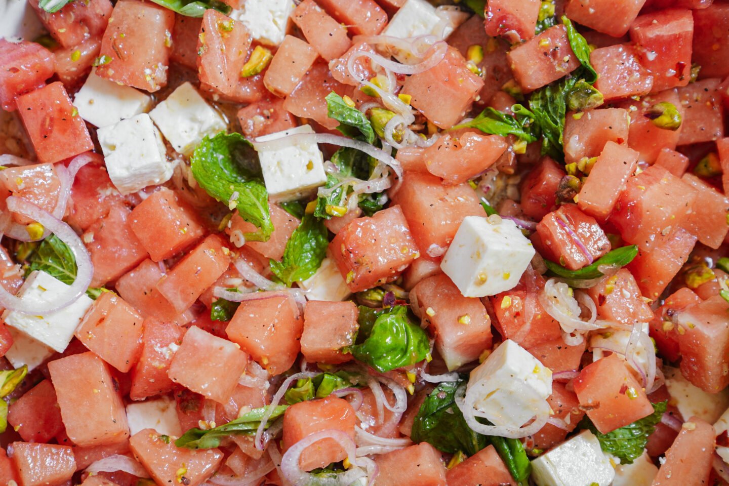 Close-up of a watermelon salad with cubes of watermelon and feta cheese, thinly sliced onions, fresh mint leaves, and chopped pistachios, all mixed together.