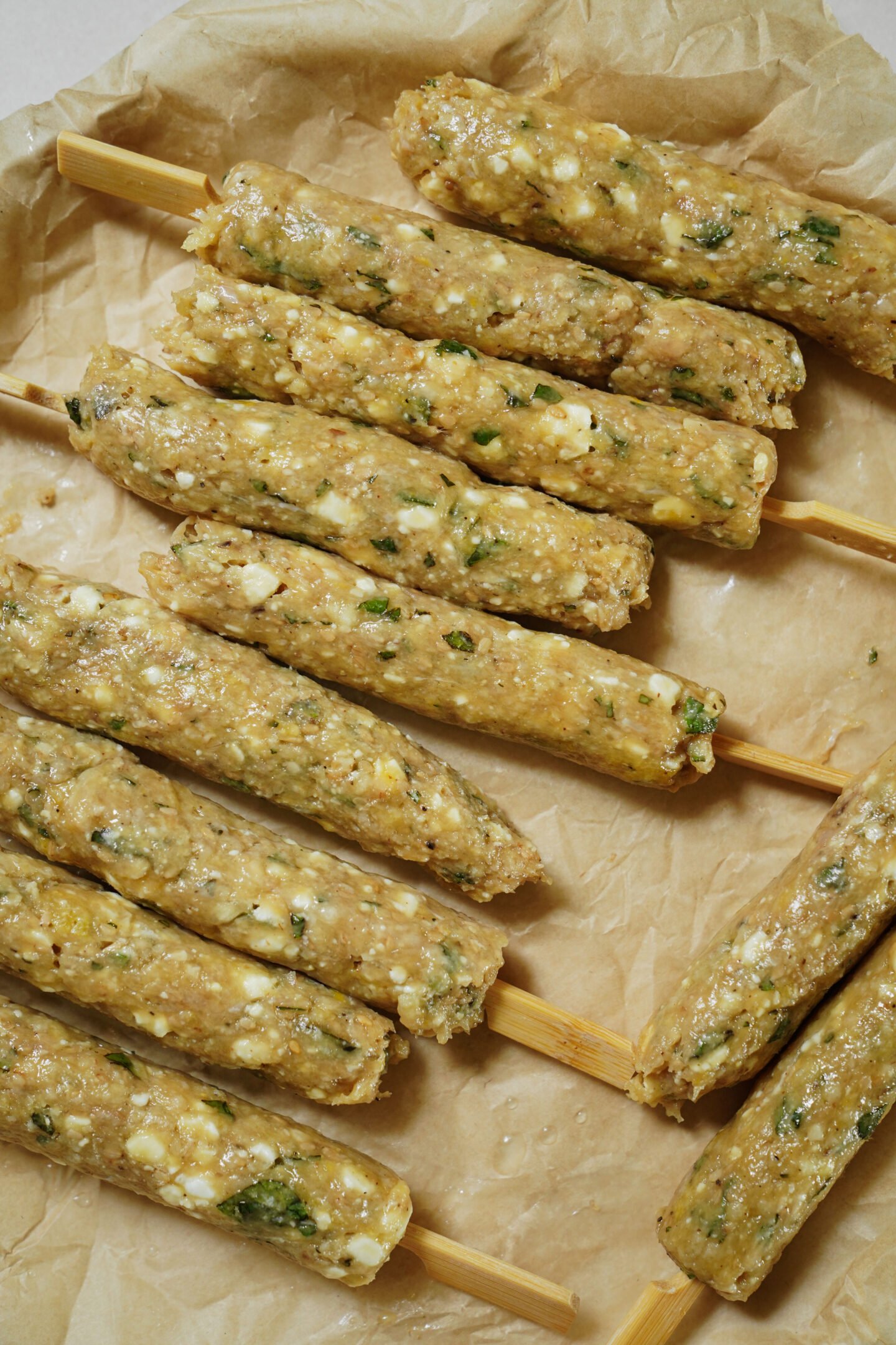 Uncooked kebabs made of ground meat, herbs, and cheese are shaped onto wooden skewers and arranged on brown parchment paper.
