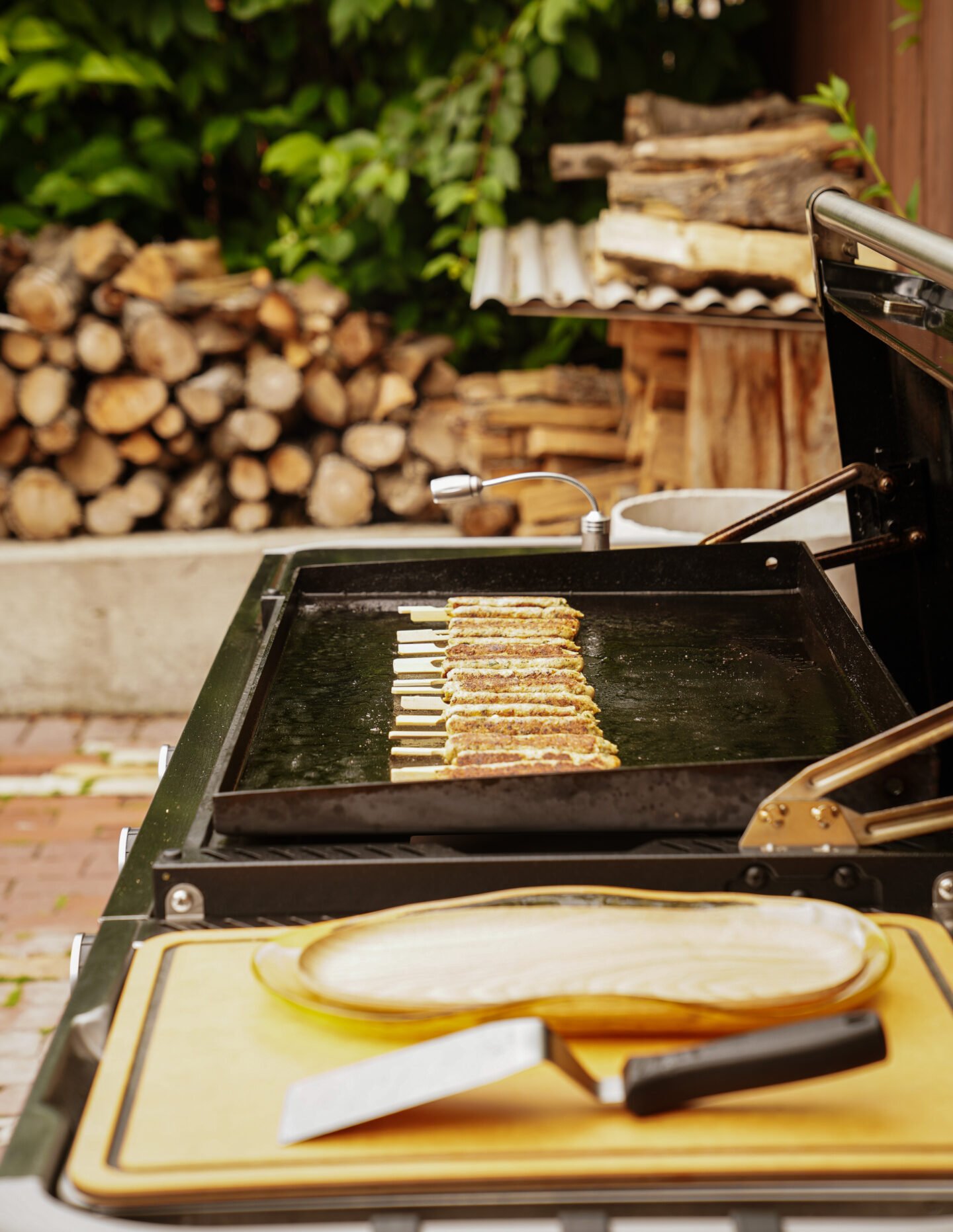 Meat skewers cooking on an outdoor grill with a wooden cutting board and knife in the foreground, and stacked firewood in the background.