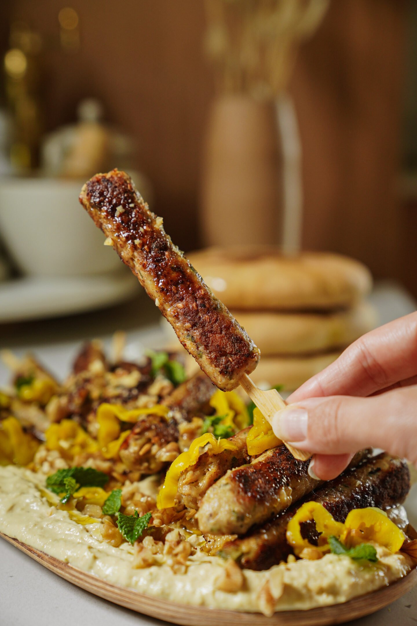 A hand holds a grilled meat skewer above a platter of kebabs, garnished with herbs and yellow peppers, served on creamy hummus. Blurred background includes bread and tableware.