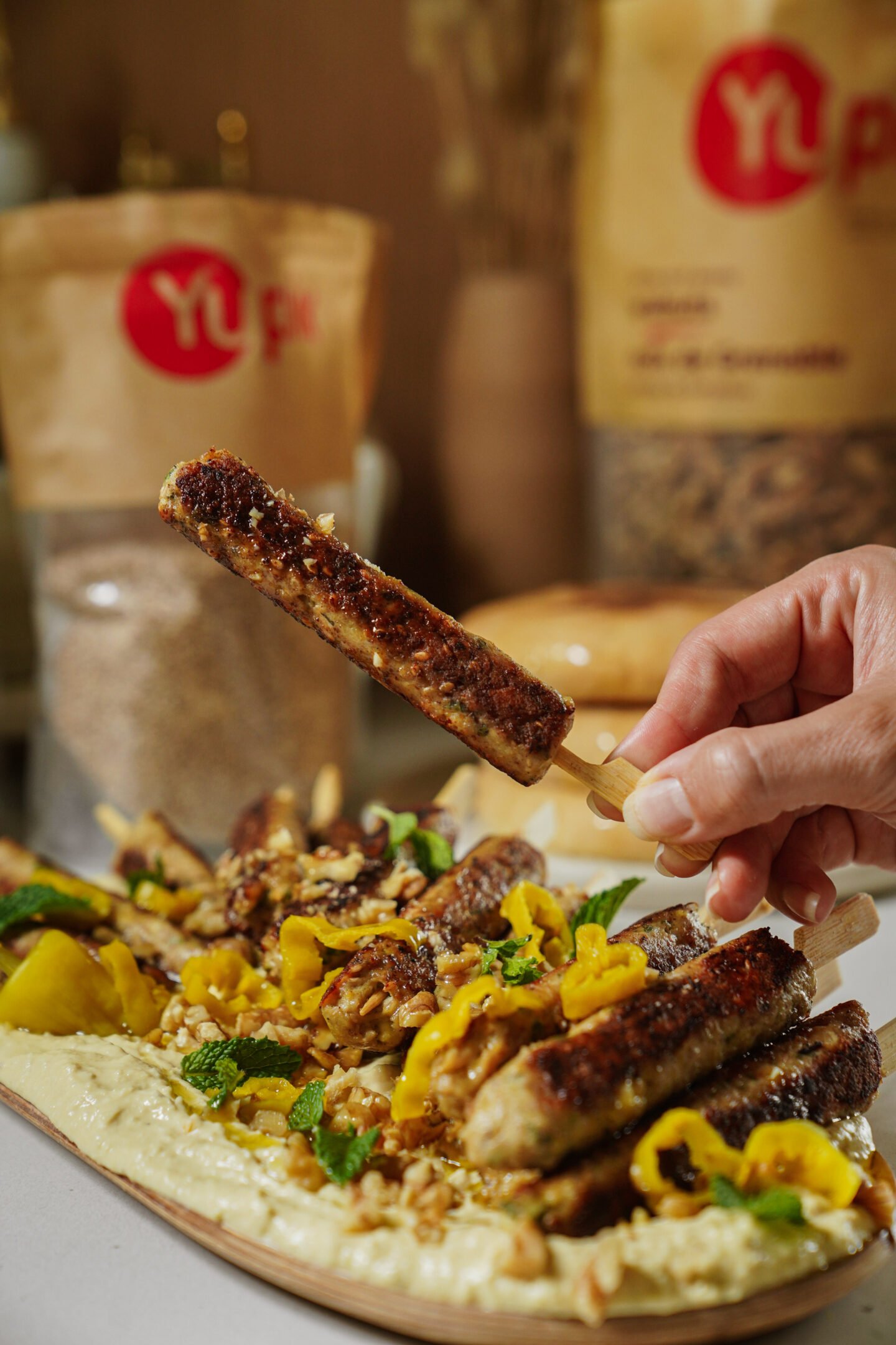 A hand holds a grilled kebab skewer above a plate of kebabs, hummus, pickled peppers, fresh herbs, and nuts. In the background, there are two large brown bags with red YL logos.