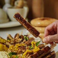 A hand holds a grilled meat skewer above a plate filled with more skewers, yellow peppers, chopped nuts, mint leaves, and a creamy dip, with bread and dishes blurred in the background.