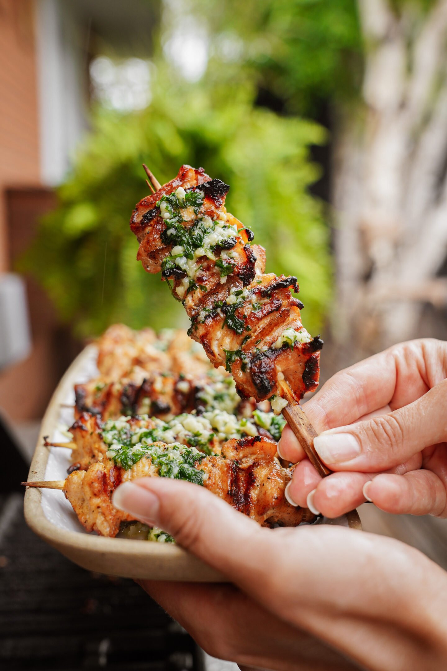 A hand holds a grilled chicken skewer topped with a green herb sauce, with more skewers on a plate in the background and greenery visible outdoors.