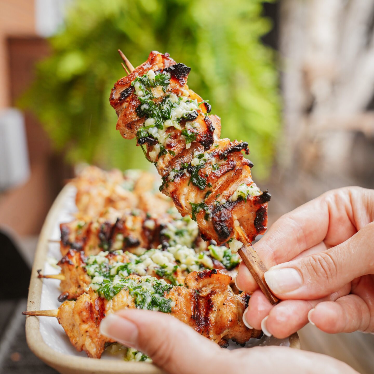 A close-up of a hand holding a grilled chicken skewer topped with green herb sauce, with more skewers on a plate in the background.