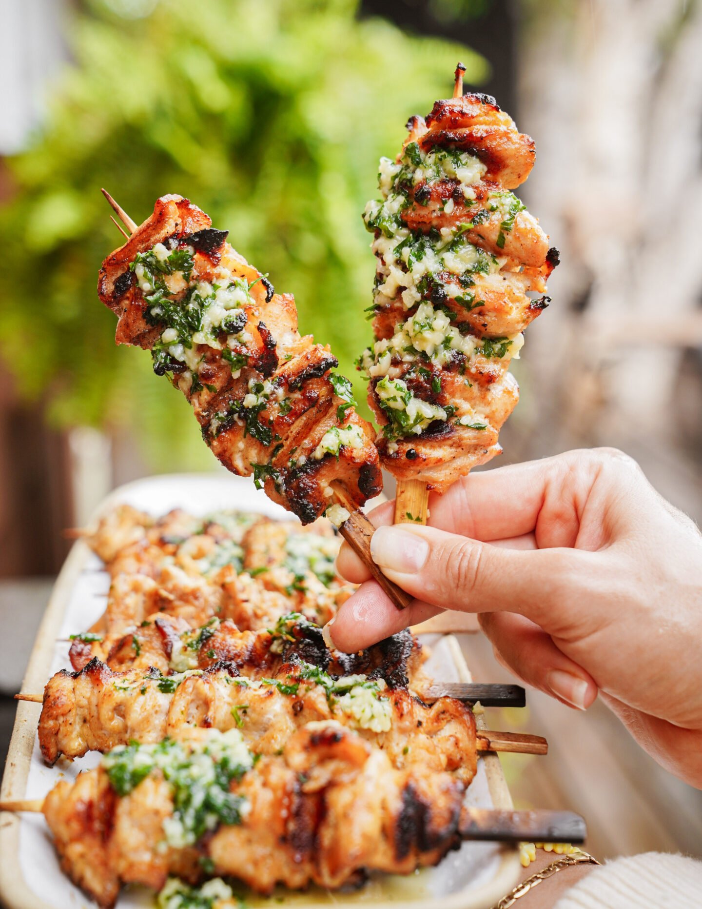 A hand holds two grilled chicken skewers topped with a green herb sauce, above a plate filled with more skewers. The background features blurred greenery.