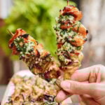 A hand holds two grilled chicken skewers topped with a chunky green herb and garlic sauce, with more skewers on a platter in the background and greenery out of focus behind.