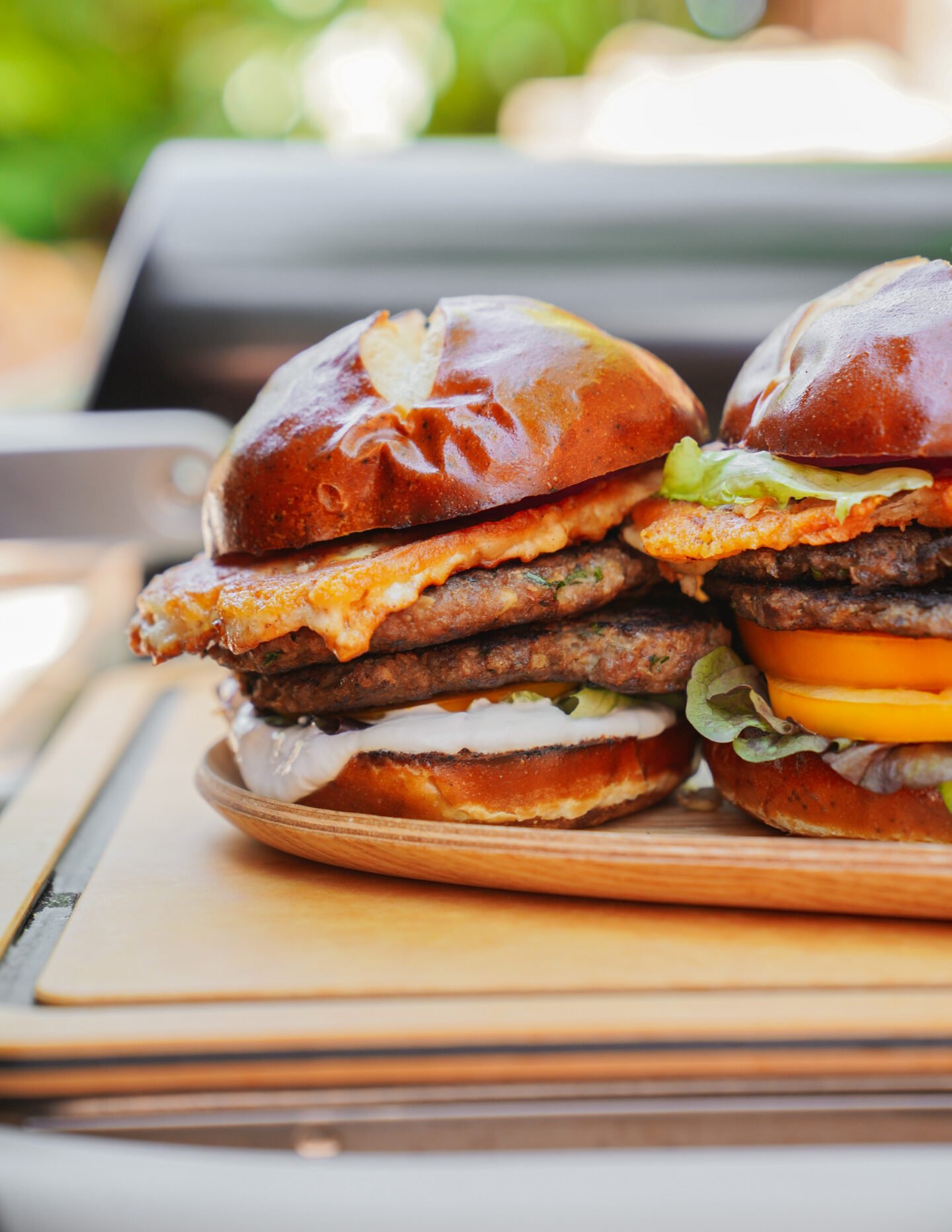Two stacked cheeseburgers with lettuce, tomato, cheese, and sauce on pretzel buns, set on a wooden platter in front of a blurred outdoor grill.