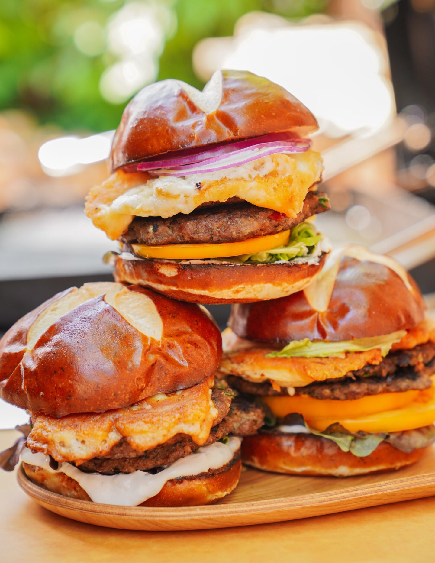 Three gourmet burgers on pretzel buns are stacked together. Each burger features a beef patty, cheese, lettuce, red onion, and a fried egg, with fresh vegetables visible in the vibrant background.