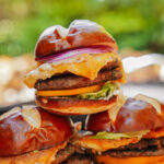 A close-up of stacked cheeseburgers on pretzel buns with melted cheese, lettuce, red onion, and beef patties, set outdoors with a blurred green background.