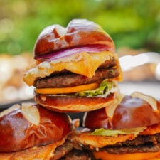 A close-up of stacked cheeseburgers on pretzel buns with melted cheese, lettuce, red onion, and beef patties, set outdoors with a blurred green background.