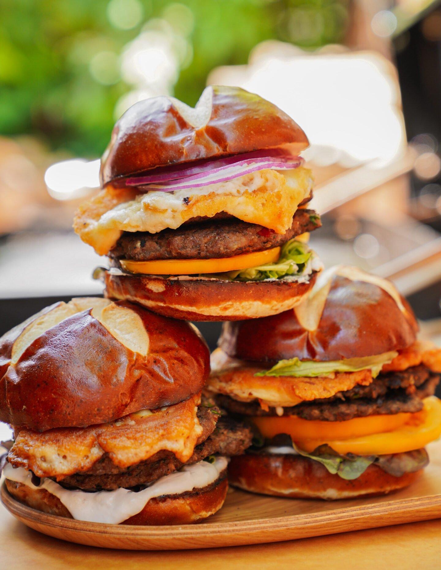 Three burgers with pretzel buns, stacked with lettuce, tomato, cheese, a fried egg, onion, and beef patties, are arranged on a wooden tray with a blurred green outdoor background.