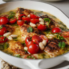 A baked chicken dish topped with cherry tomatoes, white beans, fresh herbs, and a golden sauce, served in a white casserole dish.