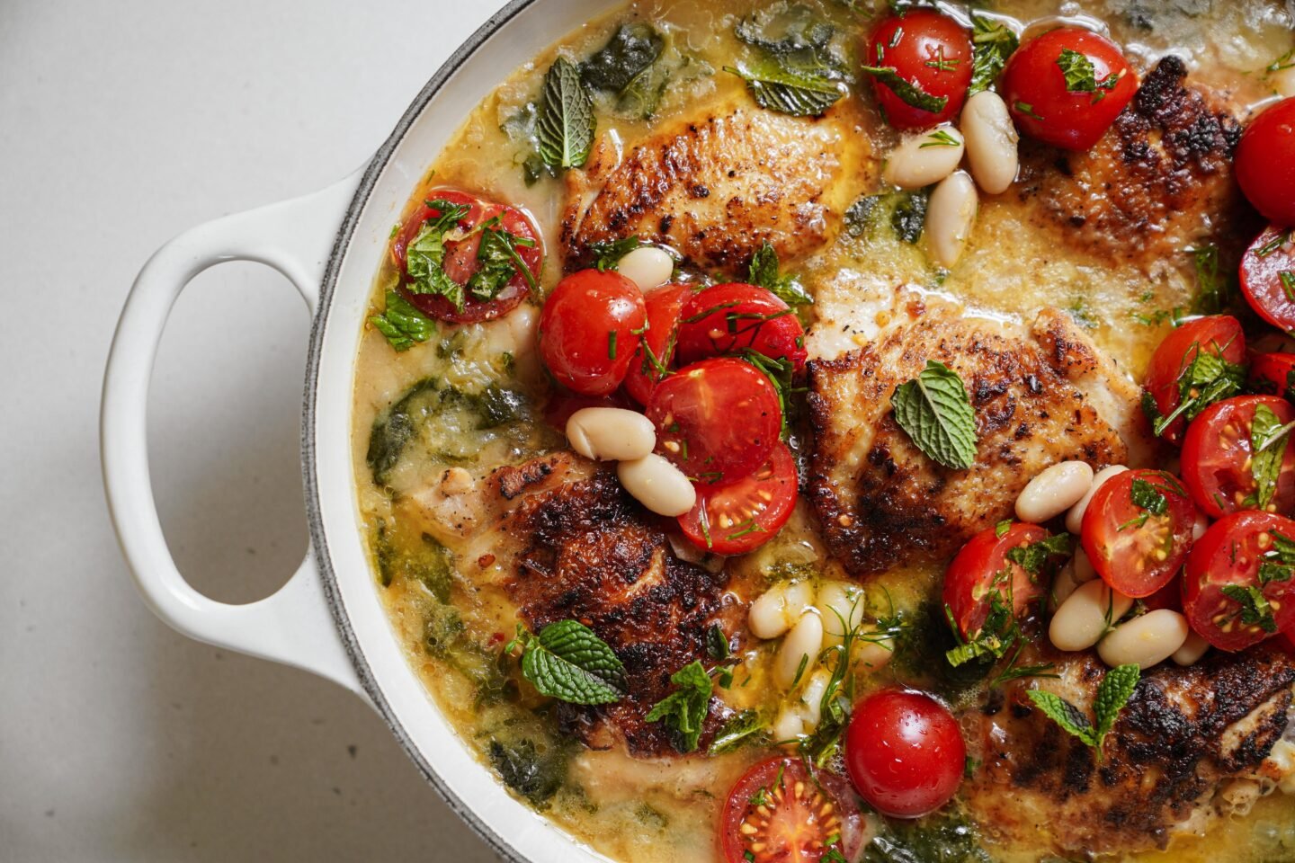 A white pan filled with browned chicken thighs, cherry tomatoes, white beans, spinach, and fresh mint leaves in a light, creamy sauce.