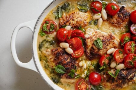 A white pan filled with browned chicken thighs, cherry tomatoes, white beans, spinach, and fresh mint leaves in a light, creamy sauce.