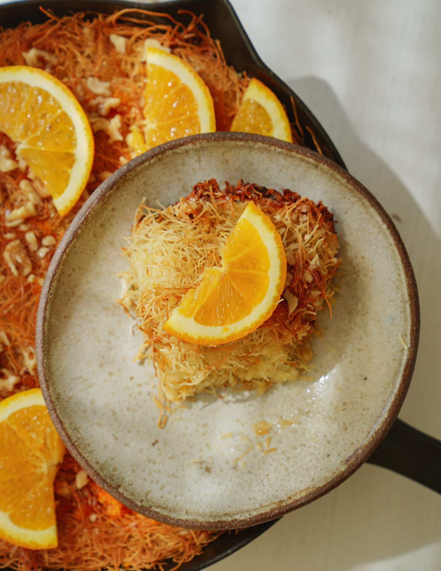 A serving of baked shredded pastry topped with an orange slice sits on a beige plate, with more pastry and orange slices visible in a dark skillet beside it.
