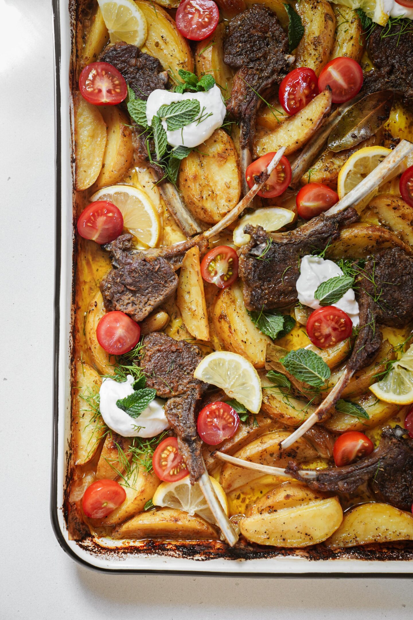 A sheet pan filled with roasted lamb chops, golden potato wedges, lemon slices, cherry tomato halves, fresh herbs, and dollops of yogurt, all arranged on a white background.