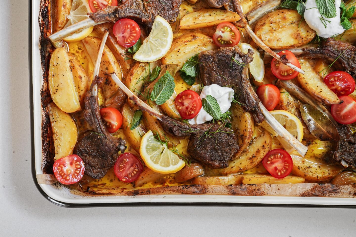 A tray of roasted lamb chops with golden potato wedges, cherry tomato halves, lemon slices, fresh mint leaves, and dollops of white sauce, all garnished and ready to serve.