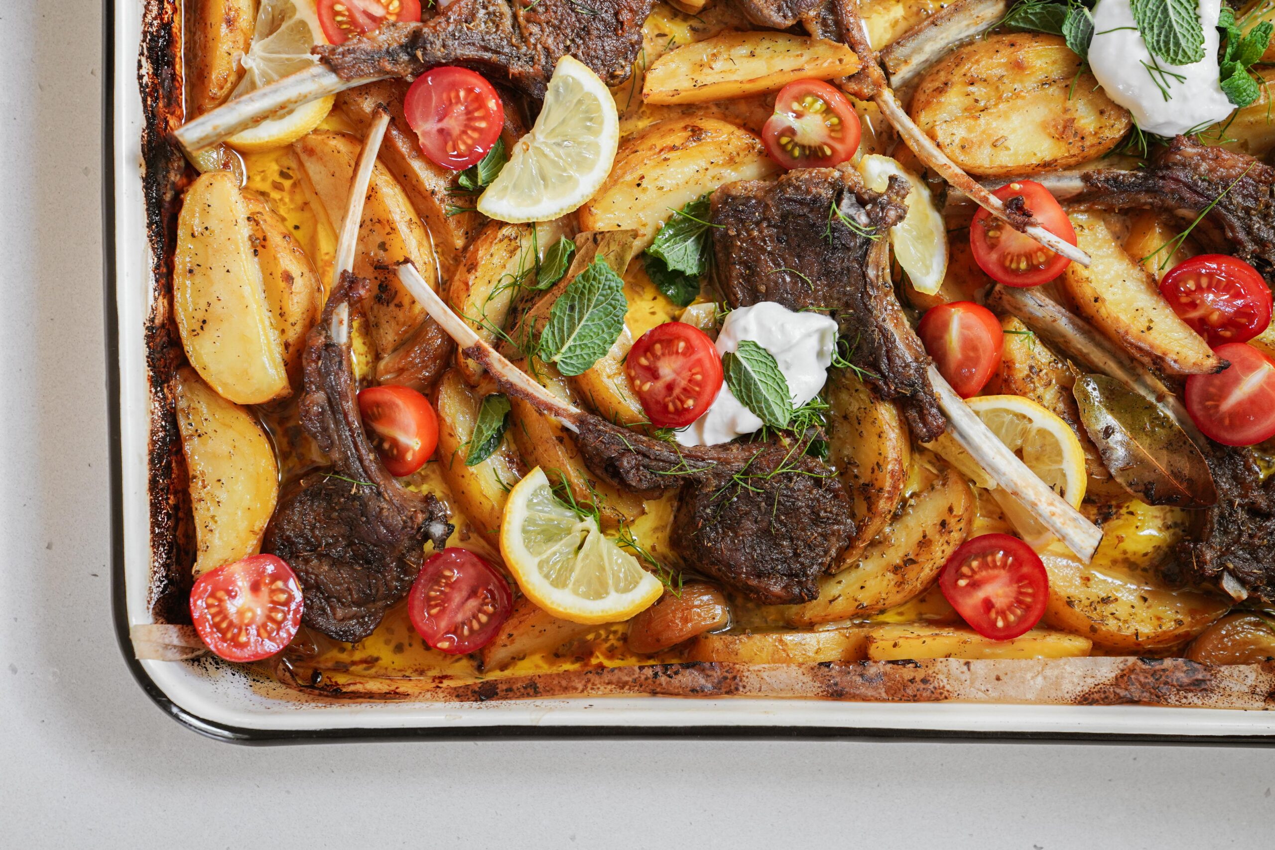 A tray of roasted lamb chops with golden potato wedges, cherry tomato halves, lemon slices, fresh mint leaves, and dollops of white sauce, all garnished and ready to serve.