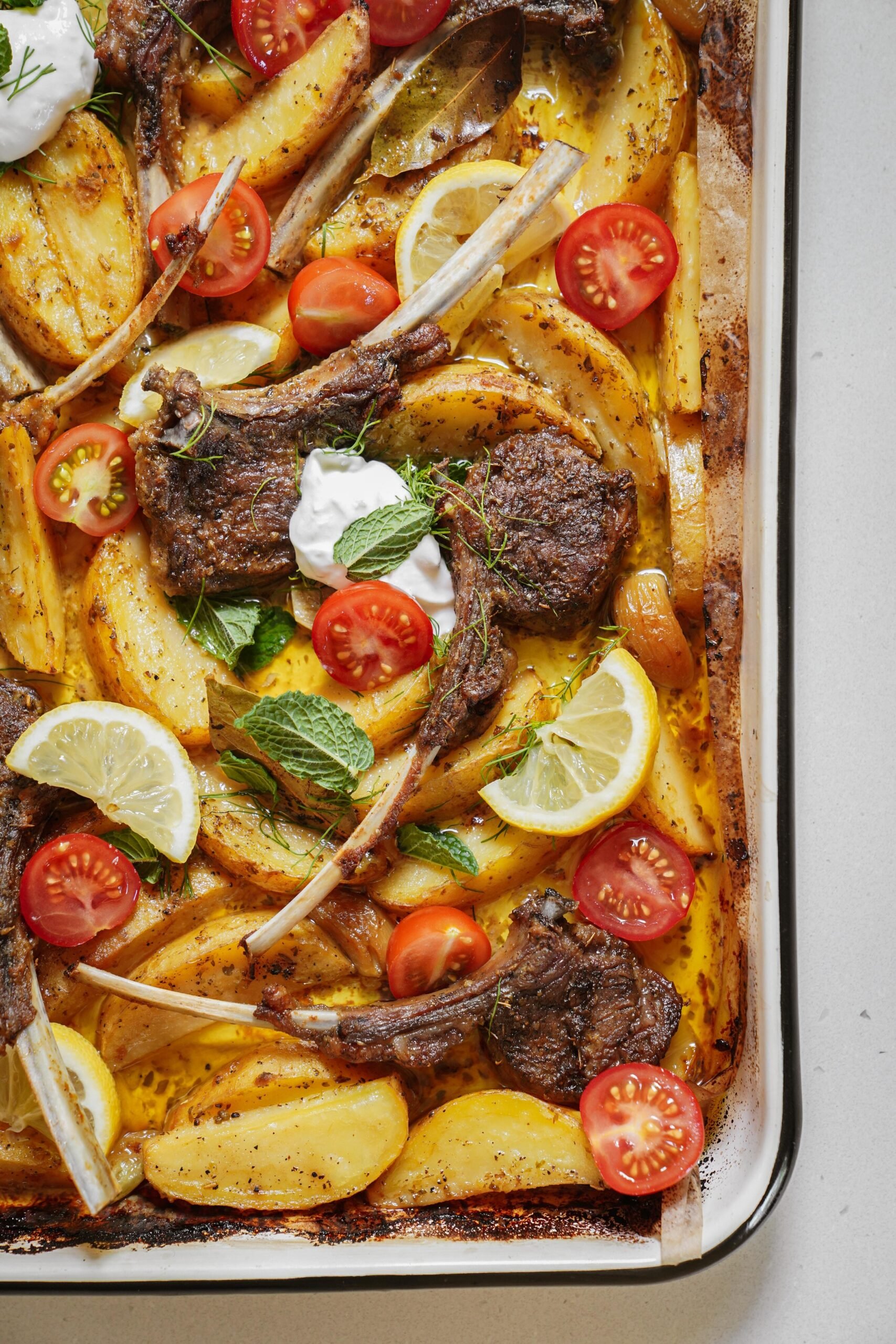 A baked tray with lamb chops, golden potato wedges, cherry tomato halves, lemon slices, and dollops of white sauce, garnished with fresh mint and herbs.