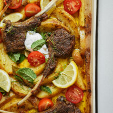 Lamb chops with roasted potato wedges, lemon slices, cherry tomato halves, fresh mint, and a dollop of sauce on a baking tray. The dish is colorful and garnished with herbs.