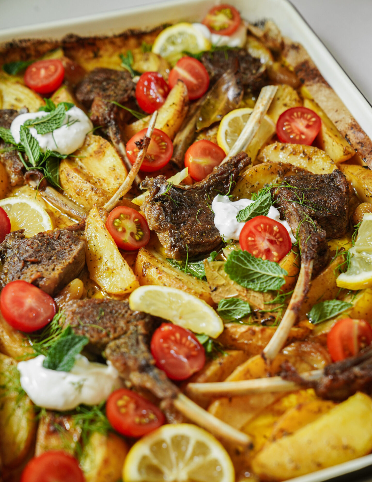 A tray of roasted lamb chops with potato wedges, cherry tomatoes, lemon slices, fresh mint leaves, and dollops of yogurt, all garnished and baked together.