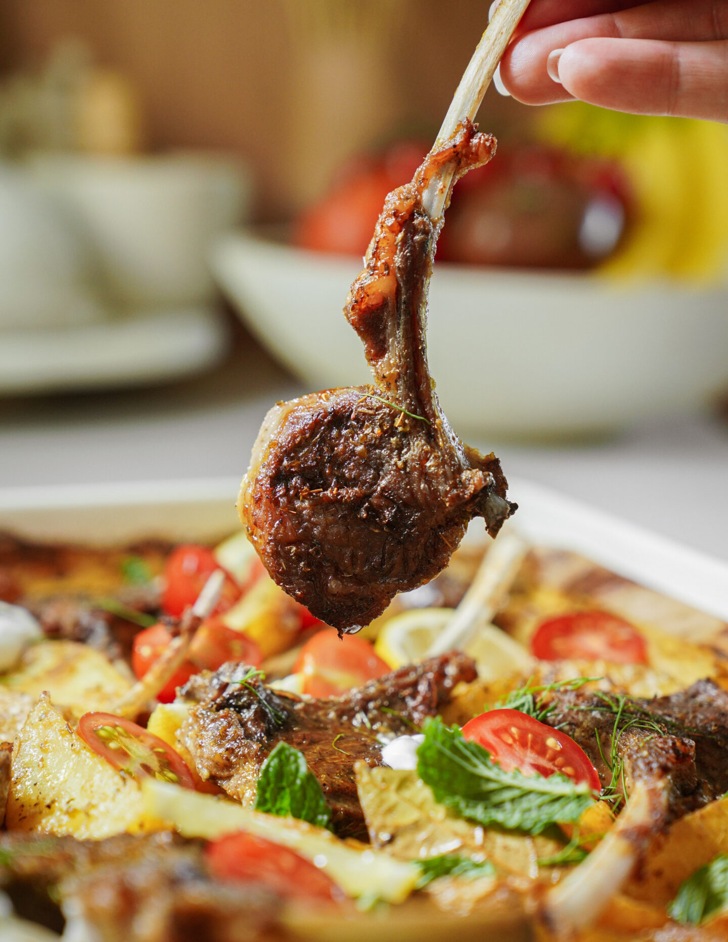 A hand holds up a cooked lamb chop above a dish filled with more lamb chops, roasted potatoes, cherry tomatoes, and fresh mint leaves. The scene is brightly lit and appetizing.