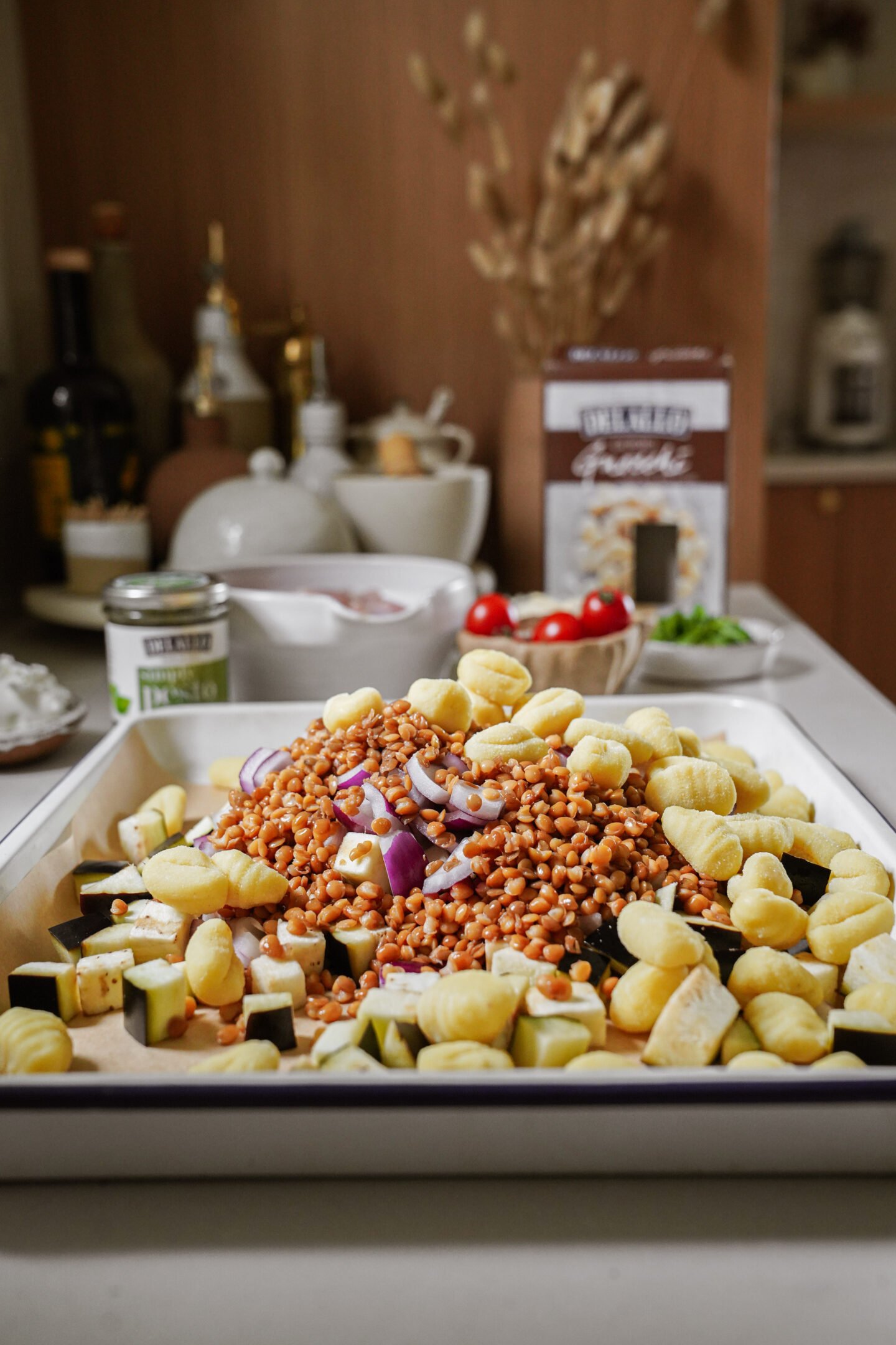 A tray filled with uncooked gnocchi, chopped zucchini, red onion, and lentils sits on a kitchen counter. Ingredients such as pesto, cherry tomatoes, and a box of gnocchi are visible in the background.