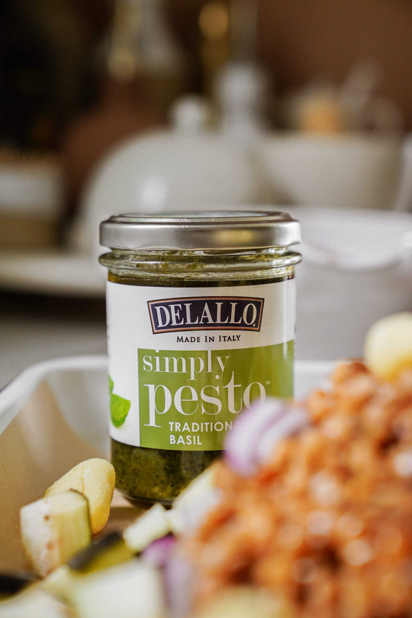 A jar of DeLallo Simply Pesto Traditional Basil is displayed on a table, with blurred food items and kitchenware in the background.