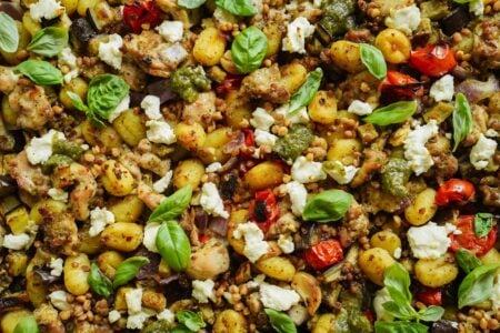 A close-up of baked gnocchi with cherry tomatoes, red onions, crumbled feta, basil leaves, chicken, pesto, and pine nuts, all mixed and topped with herbs.