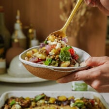 A hand holds a ceramic bowl of roasted vegetable salad with feta, lentils, and fresh basil as a gold spoon scoops up a bite; a baking dish and kitchen items are blurred in the background.