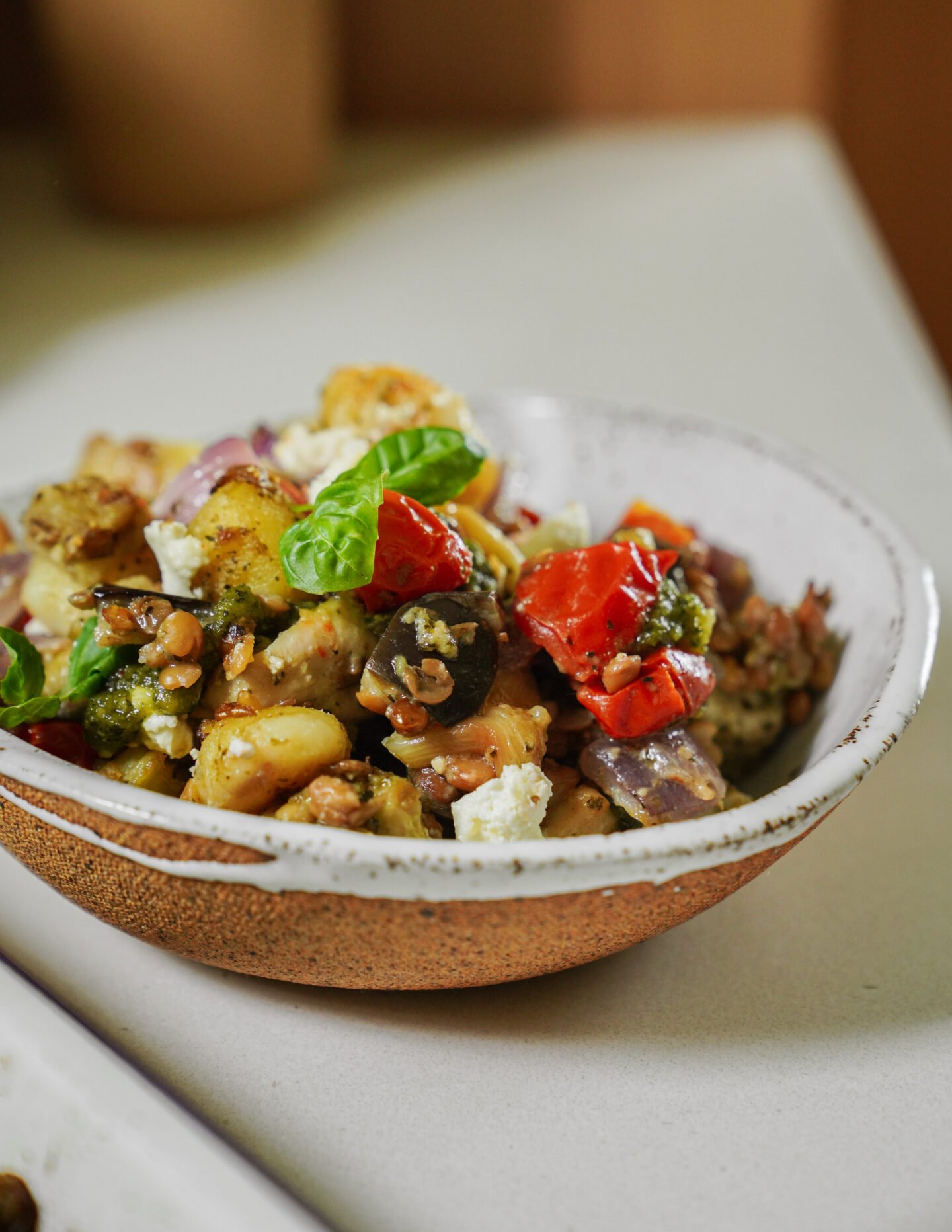 A rustic ceramic bowl filled with a colorful salad containing gnocchi, cherry tomatoes, olives, red onions, fresh basil, and crumbled cheese, set on a light countertop.