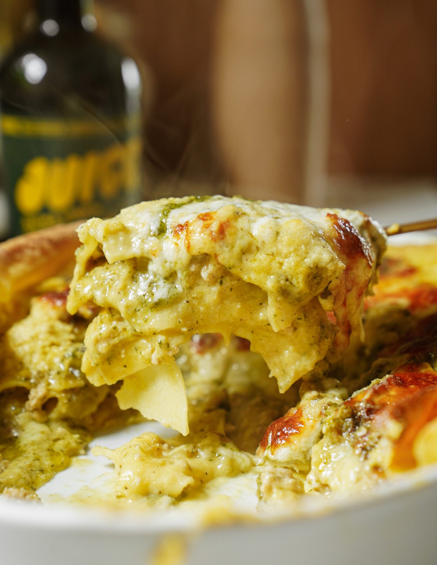 A close-up of a creamy, cheesy pasta bake being served from a white dish, with melty cheese and green pesto sauce. A blurred bottle is visible in the background.