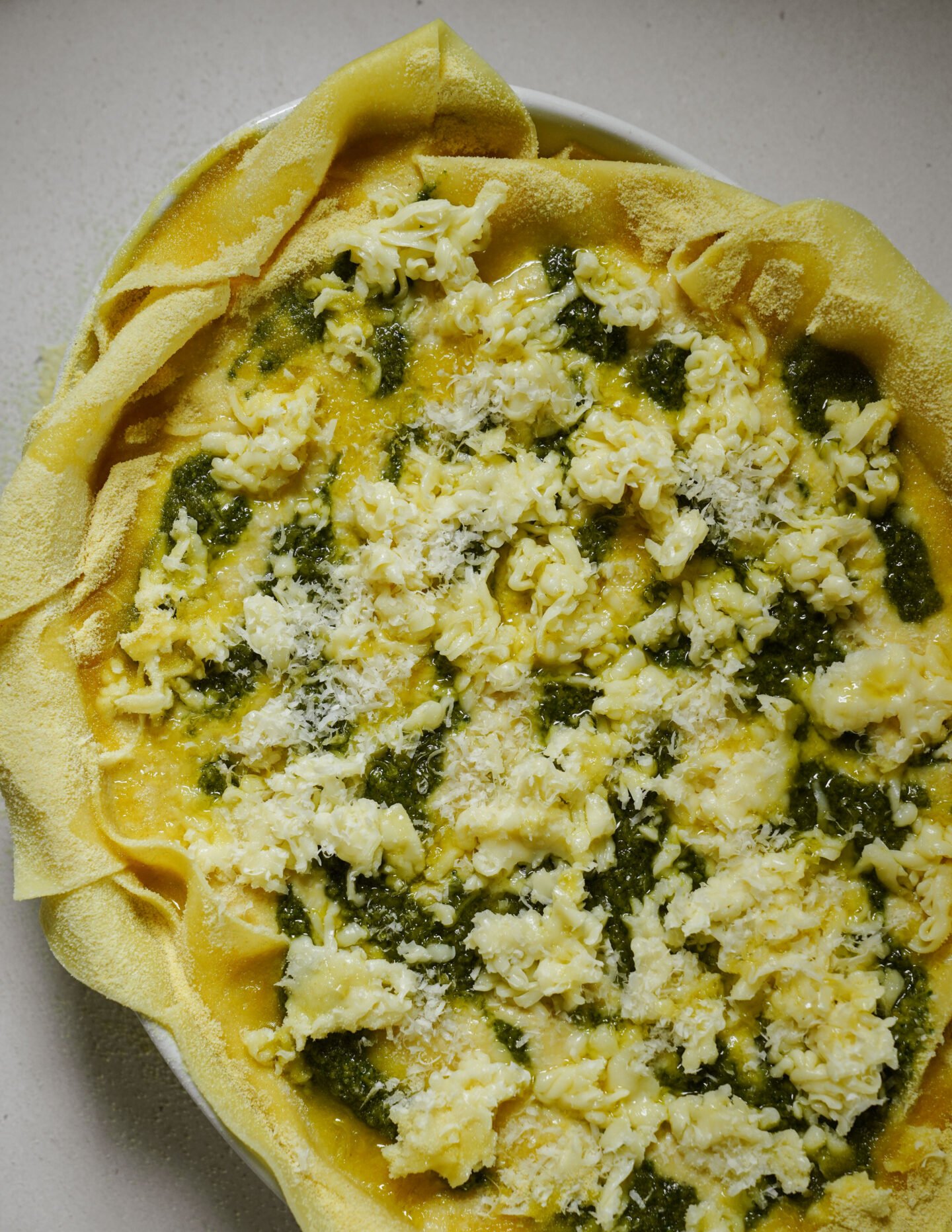 A close-up of an unbaked lasagna in a white dish, featuring layers of pasta sheets, green pesto, grated cheese, and a light dusting of shredded Parmesan on top.