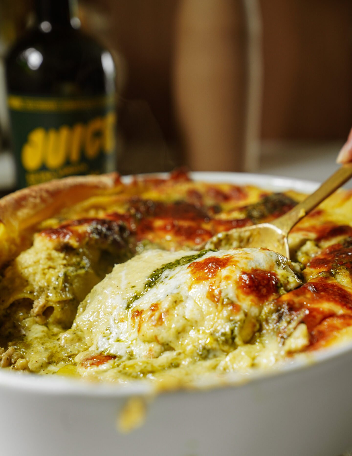A close-up of a cheesy baked pasta dish with golden melted cheese and green pesto, being served with a spoon. A bottle with a yellow “JUICE” label is blurred in the background.