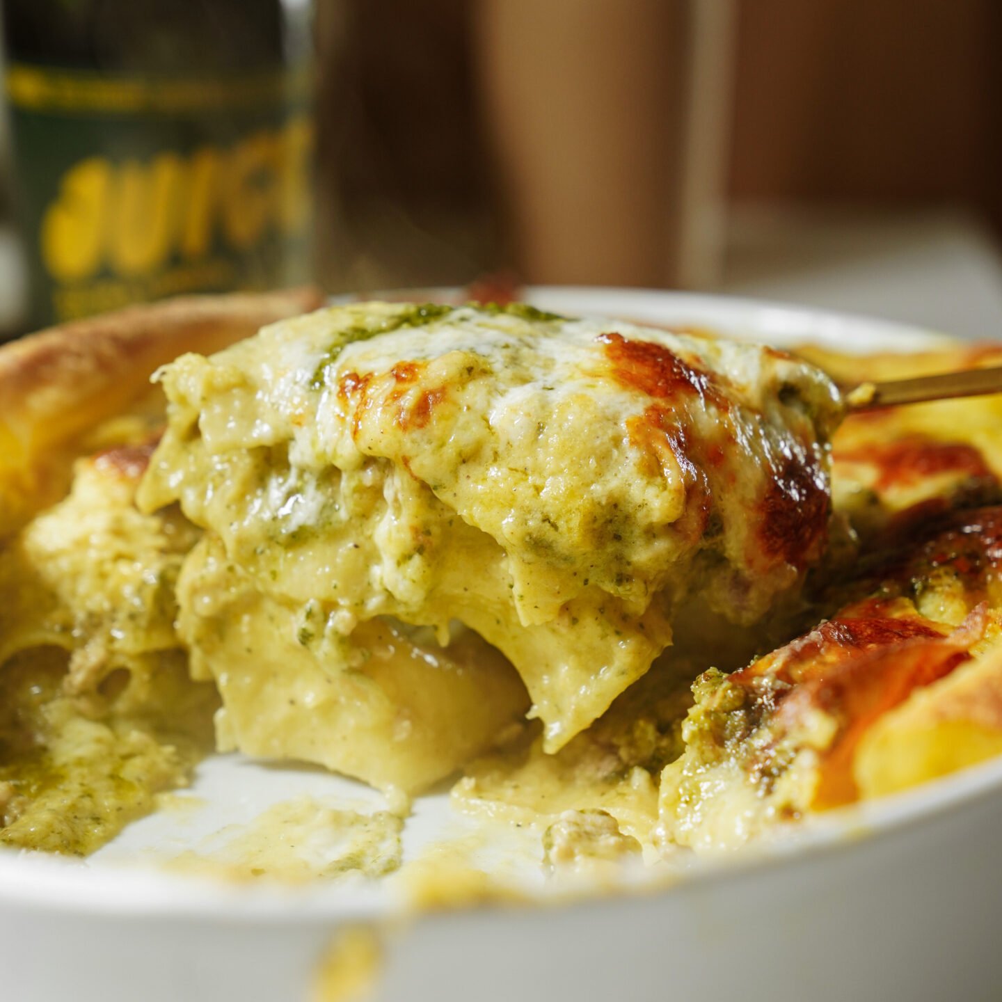 A close-up of a golden, cheesy baked pasta dish being served with a spoon, showing layers of pasta, creamy sauce, and green pesto in a white baking dish.