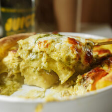 A close-up of a golden, cheesy baked pasta dish being served with a spoon, showing layers of pasta, creamy sauce, and green pesto in a white baking dish.